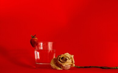 Close-up of a glass and strawberry resting atop and a white rose on a red background