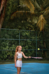 A beautiful girl with long blond hair gathered in a ponytail plays tennis. Tennis court. A girl holds a tennis racket and a tennis ball. A game. Sport. Tennis court on the island, among palm trees.
