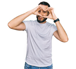 Fototapeta premium Young man with beard wearing casual white t shirt doing heart shape with hand and fingers smiling looking through sign