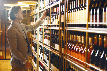 Sommelier stands in cellar and taking out one wine of many submitted from wooden shelf. Male...