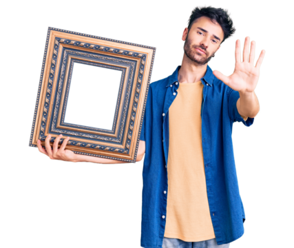 Young hispanic man holding empty frame with open hand doing stop sign with serious and confident expression, defense gesture - Powered by Adobe