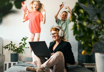 Remote work at home: A girl in a jacket and pajamas, a call worker contacts a client via video conference. Children run around, make loud noises and get in the way. - obrazy, fototapety, plakaty