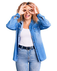Young blonde woman with curly hair wearing casual denim jacket doing ok gesture like binoculars sticking tongue out, eyes looking through fingers. crazy expression.