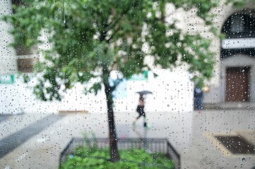 Closeup of a window covered with raindrops against a woman walking on the street with an umbrella