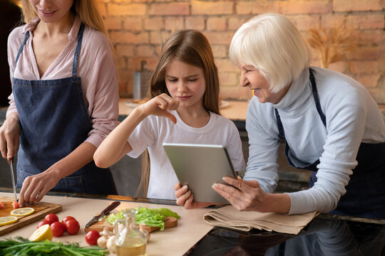 Close shot of little girl that don't like what grandmother showing her in the tablet in kitchen. Mother cutting pepper for fresh salad. Granny with granddaughter choosing what cook to internet - Powered by Adobe