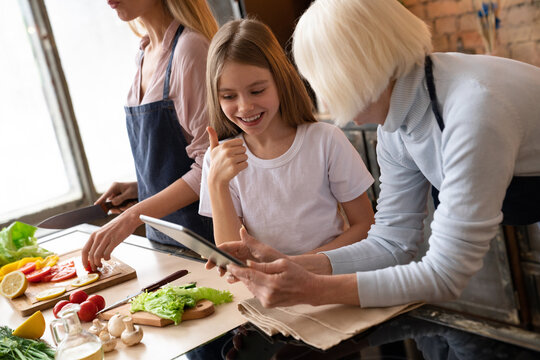 Side view shot of smiling girl that like what grandmother showing her in the tablet in kitchen. Cooking young woman on the background. Granny with granddaughter choosing what cook to internet