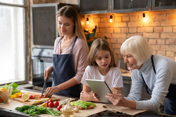 Horizontal shot of senior woman that looking in the tablet with her granddaughter and smiling. Mother cooking vegetarian dinner in kitchen. Women searching recipe in culinary website