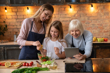 Beautiful young woman squeezing lemon in fresh vegetable salad with her little cute daughter. Grandmother reading recipe for vegetarian vegan healthy food. Happy family concept