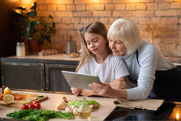 Elder woman with her little granddaughter looking in the tablet while cooking vegetables and fruits in home kitchen. White hair grandmother and cute girl watching to screen pad