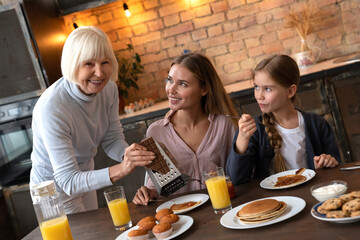 Side view of cheerful mature woman rubbing chocolate in the plate with pancakes for her daughter....