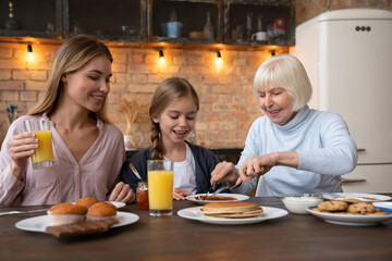 Happy mom, daughter and grandmother eating breakfast in kitchen. Family concept. Careful granny cutting pancakes for her little granddaughter, attractive woman watching for them