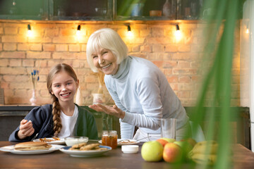 Front shot of happy little girl having fun with her senior grandmother on the kitchen sitting at...