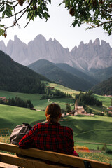 Woman traveler sitting on bench in checkered shirt and looking at amazing Dolomites Mountains. Wanderlust travel concept, atmospheric epic moment, Santa Magdalena, Italy. - obrazy, fototapety, plakaty