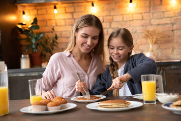 Smiling little girl sharing a piece of pancake on plate with her mother at breakfast in modern kitchen. Young pretty woman have lunch time with her cute daughter. Happy family concept