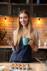 Vertical shot of cheerful attractive woman chilling in kitchen while cooking looking at camera. Beautiful young woman with cup of tea listening music on mobile phone in kitchen