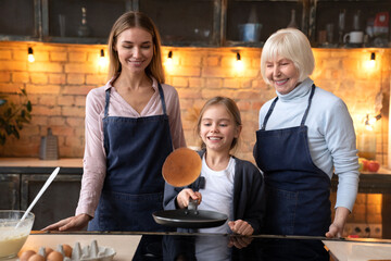 Happy little girl cooking healthy breakfast in kitchen with her mother and grandmother. Young attractive mother and granny smiling and looking on flying pancake