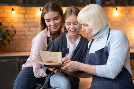 Happy three generations of women reading together recipe book in kitchen while cooking at home. Grandmother show her beautiful daughter and little grandchild secret family recipe