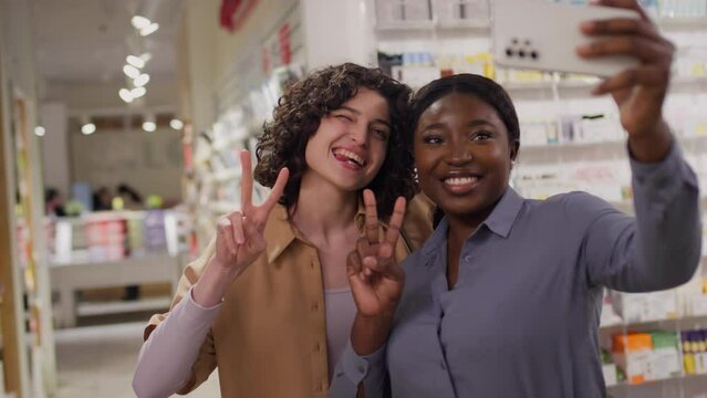Medium shot of young Caucasian and African American female friends taking selfie photos on smartphone together in cosmetics store, smiling, showing fun hand gestures and making faces