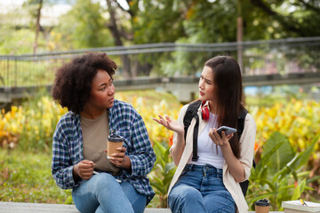 Two women sitting on a bench in a park, one of them holding a cup. They are talking to each other - Powered by Adobe