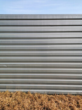 A textured background with a metal gray fence with horizontal stripes - stiffeners, in front of which is dry yellow beige grass, and behind them is a light blue sky