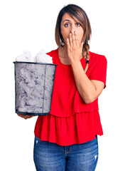 Young beautiful woman holding paper bin full of crumpled papers covering mouth with hand, shocked...
