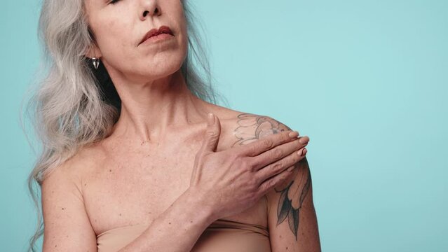 A mature woman with silver hair and a floral tattoo is seen applying cream to her shoulder, suggesting skincare and self-care routines.