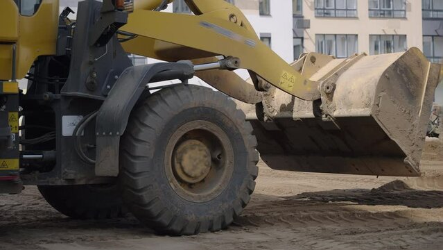 A construction site is being cleared by a yellow bulldozer with strong tires, moving dirt and debris. The vehicles wheels are covered in mud and tread marks are visible on the asphalt