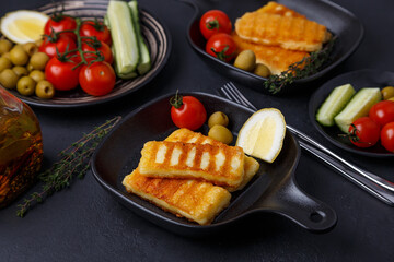 Grilled halloumi cheese in a black pan with olives, tomatoes, cucumbers, thyme and lemon. Traditional cheese of the Eastern Mediterranean. Black background, selective focus, close-up.