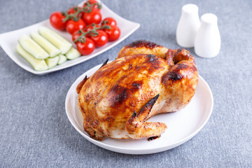 Chicken baked whole to a delicious crust. A traditional dish. In the background are cucumbers and cherry tomatoes on a white plate. Close-up, selective focus, gray background.