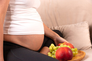 A pregnant woman sits on the couch with a plate of fruit.