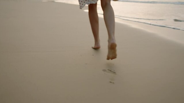 Female feet running barefoot on tropical sandy beach at summer sunset. Slim woman's legs run on sand, leaving footprints. Pretty girl in white dress, carrying straw hat at the seaside surf. Low angle.