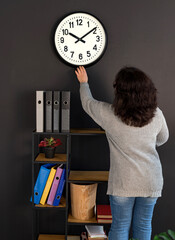 A woman in an office is facing a large wall clock. Next to her is a bookshelf with folders of...