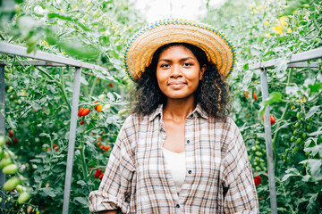 Amidst tomato plants a cute farmer woman stands smiling in her greenhouse. Her occupation is evident in the portrait depicting care and growth in the business of agriculture. Working outdoors is.