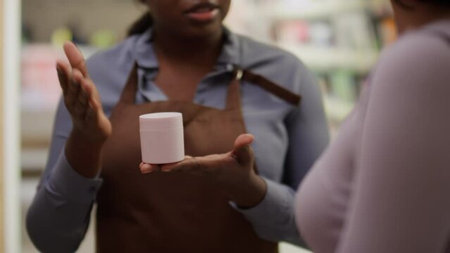 Close-up footage of hands and midsection of unrecognizable dark-skinned female sales assistant holding jar of cream and advertising its benefits to anonymous woman shopping for makeup in store