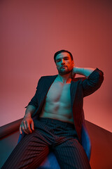 man with bare chest posing in elegant pinstripe suit and sitting in studio with red and blue light