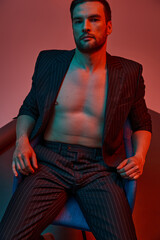 brunette man with bare chest posing in pinstripe suit and sitting in studio with red and blue light