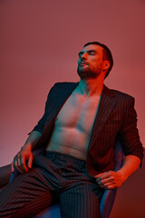 handsome man with bare chest posing in pinstripe suit and sitting in studio with red and blue light