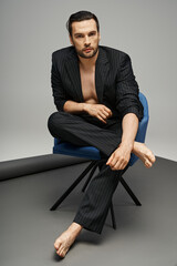 striking pose on handsome man with  bare chest posing in pinstripe suit on armchair on grey backdrop