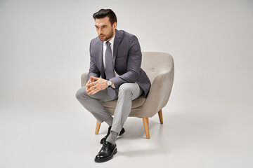 confident and handsome businessman in elegant suit sitting on comfy armchair on grey backdrop