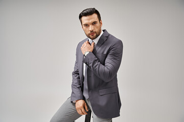 handsome corporate man in elegant formal wear leaning on high stool on grey background, business