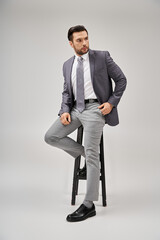 handsome and confident businessman in formal wear leaning on high stool on grey background - 768021768