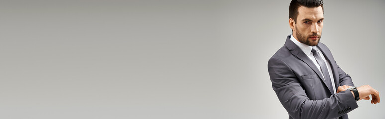 handsome businessman posing in elegant suit checking his wristwatch on grey background, banner