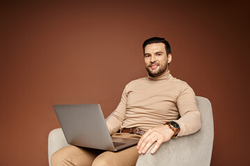 cheerful man in turtleneck sitting on comfortable armchair and using laptop on beige backdrop