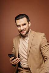 Stylish and handsome man with bristle smiling and holding smartphone on beige backdrop, social media