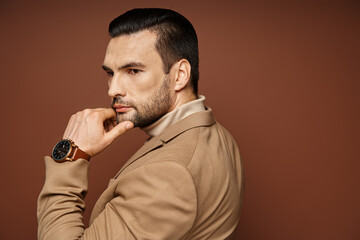 pensive man in elegant attire giving confident look while posing with hand near chin on beige
