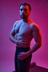 handsome man with bare chest posing in pinstripe trousers on purple backdrop with red lights