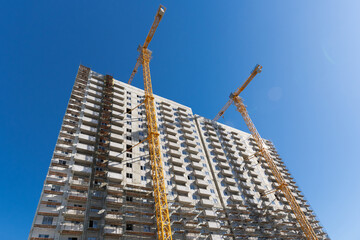 Crane and building under construction against blue sky. Construction work site. Construction of a multi-storey building using a high-rise yellow crane. Home construction.