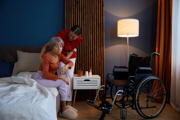 Positive home caregiver helping sad unhappy old woman in bedroom - obrazy, fototapety, plakaty