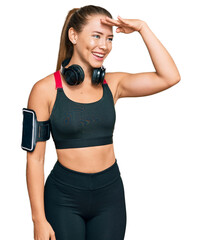 Beautiful blonde woman wearing gym clothes and using headphones very happy and smiling looking far away with hand over head. searching concept.
