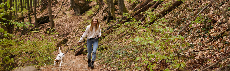 Smiling young woman in sweater and jeans walking dog on leash in forest path while hiking, banner